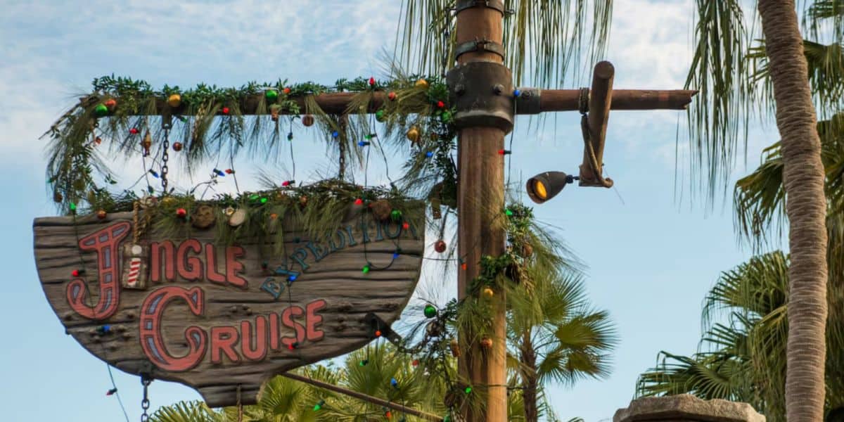 A wooden “Jingle Cruise” sign, inspired by the classic Jungle Cruise, is adorned with festive garlands and colorful holiday lights, hanging on a pole among palm trees beneath a clear blue sky.
