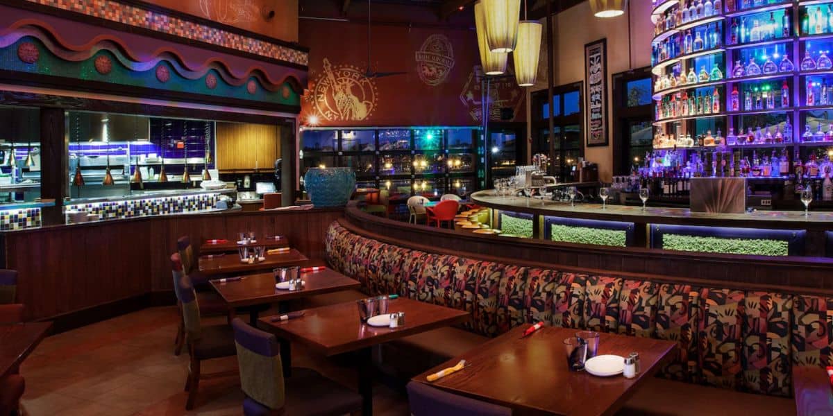 A modern restaurant interior featuring wooden tables, patterned booth seating, and a curved bar with illuminated shelves displaying various bottles. Warm lighting and decorative accents create an inviting restaurant atmosphere.
