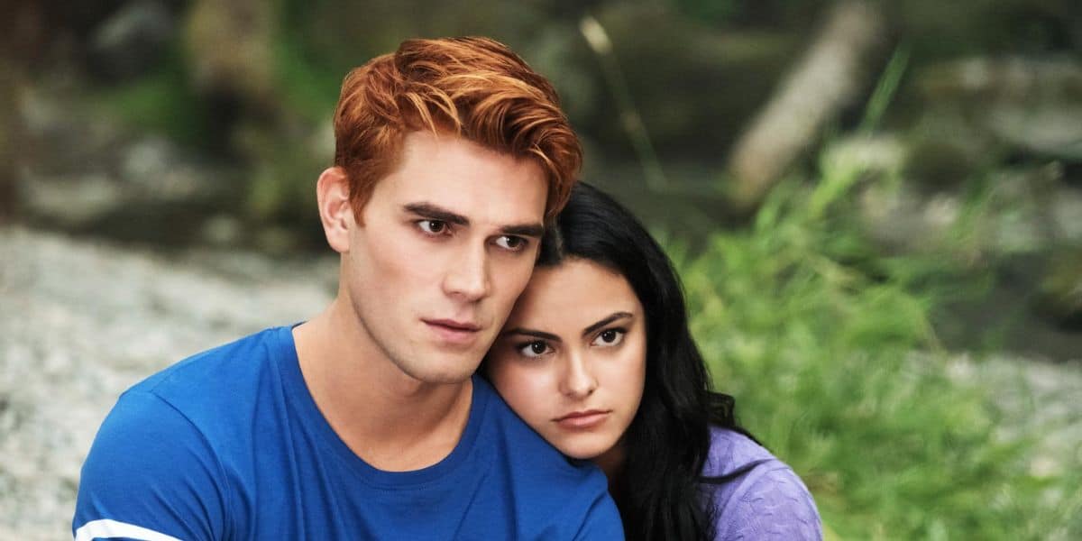 A young man with short red hair in a blue shirt sits closely with a young woman with long black hair in a purple shirt, resting her head on his shoulder. They both appear thoughtful, evoking the close bonds seen in Riverdale, with nature blurred behind them.