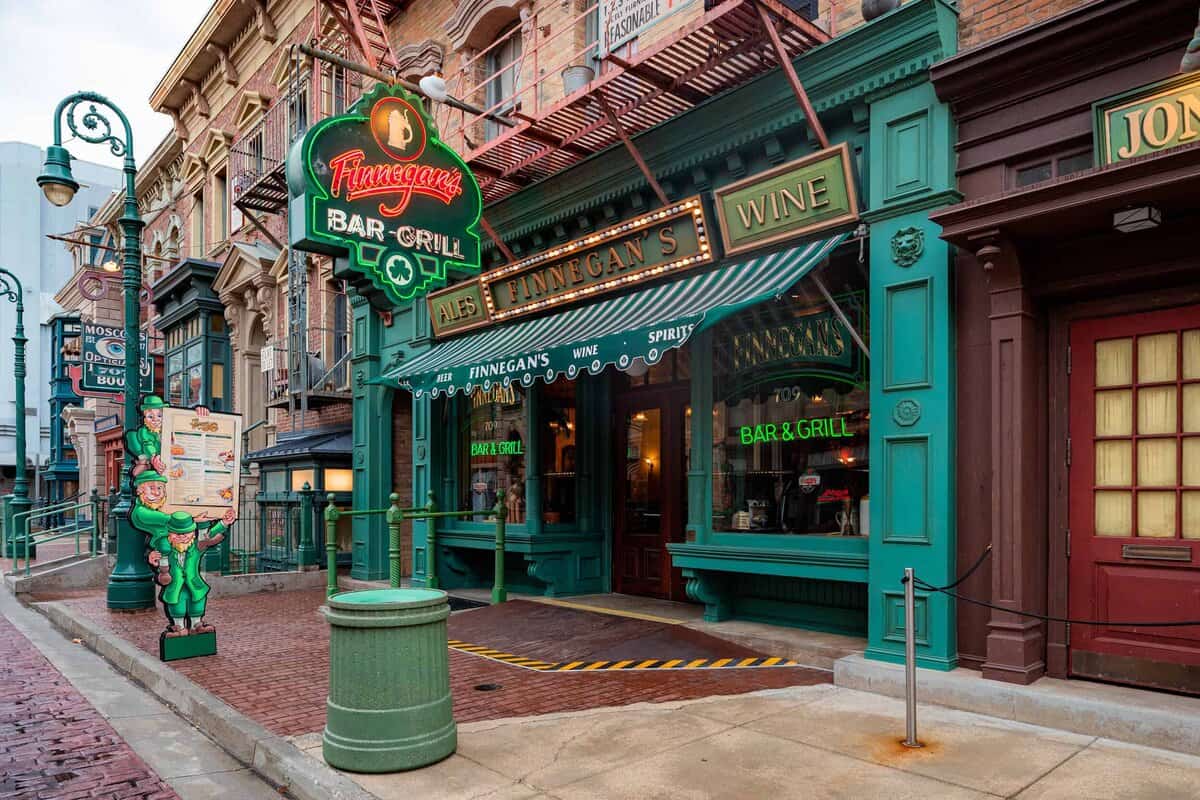 The image shows the exterior of Finnegan’s Bar & Grill, a green and red pub with neon signs and vintage-style decor, located on a brick street corner with a pergola and a cartoon leprechaun figure at the entrance.