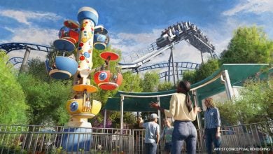 Artistโs rendering of a vibrant amusement park with a colorful spinning ride on the left, a looping roller coaster above, lush greenery, and people exploring and looking up in excitement in the foreground at this new Jurassic World themed land coming soon to this Universal park.