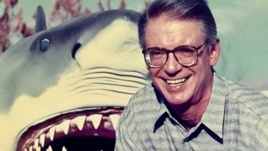 A smiling man wearing glasses and a patterned shirt poses in front of a large, open-mouthed shark model, with trees and blue sky in the background.