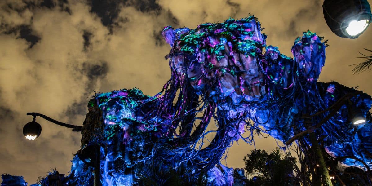 Floating mountain structures in Animal Kingdom glow with vibrant blue and purple lights at night, set against a cloudy sky. Lush plants and hanging vines enhance the fantastical, alien landscape.