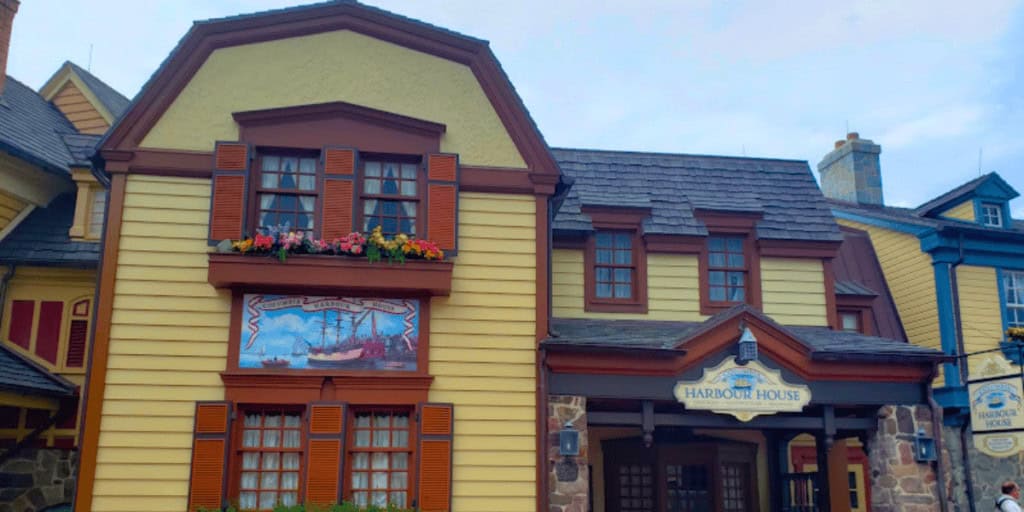 A yellow and brown colonial-style building with flower boxes, wooden shutters, signs reading “Harbour House,” and a painted mural of ships. Another similar building is partially visible on the left and right sides.