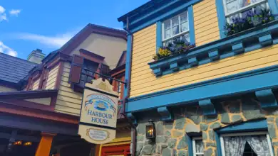 A sign for Columbia Harbour House hangs outside a yellow and blue colonial-style building with stone walls and shuttered windows decorated with flower boxes, under a blue sky with scattered clouds.