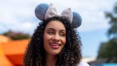 A woman with curly hair is smiling and wearing Mickey Mouse ears with a large white bow at Epcot. The background is outdoors with some blurred orange and green elements.