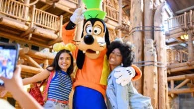 Two smiling women pose with a person in a Goofy costume inside a rustic, wooden building while someone takes their photo with a smartphone.
