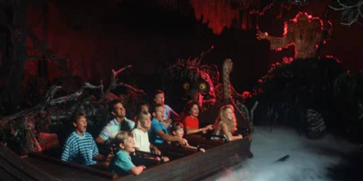 A group of people sit in a boat on a dark amusement park ride, surrounded by fog and eerie, glowing red lighting, as they drift toward a maelstrom with a large, menacing animatronic figure looming ahead.