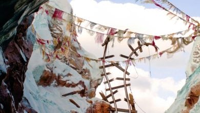 A broken, twisted railway track juts out over a snowy mountain pass near Everest, surrounded by colorful prayer flags strung between rocky, snow-covered peaks under a partly cloudy sky.