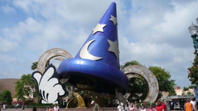 A large blue sorcerer's hat adorned with silver stars and a crescent moon, along with a giant white-gloved hand, serves as a landmark at a theme park where people stroll under a partly cloudy sky.