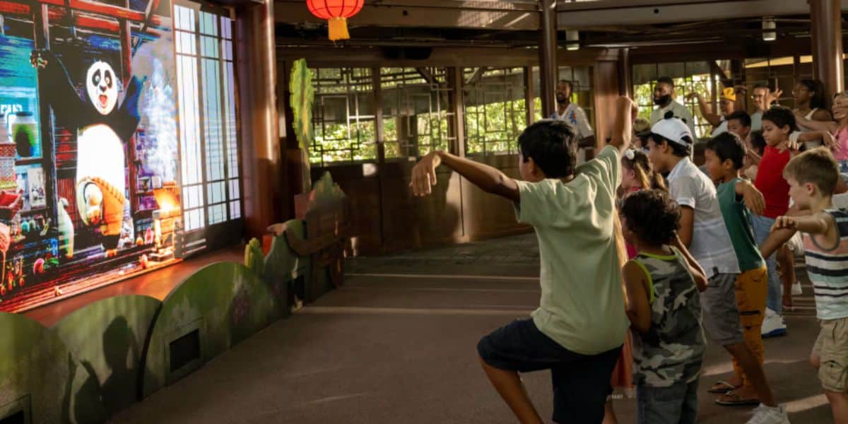 Children mimic martial arts poses in front of a large screen displaying Kung Fu Panda in an indoor play area, while adults watch in the background.