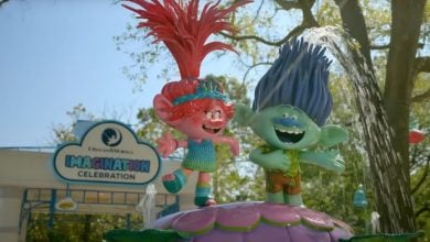 Two colorful Trolls characters stand on a flower statue with water spraying behind them, smiling joyfully. A sign in the background reads "DreamWorks Imagination Celebration." Trees and sunlight surround the scene.
