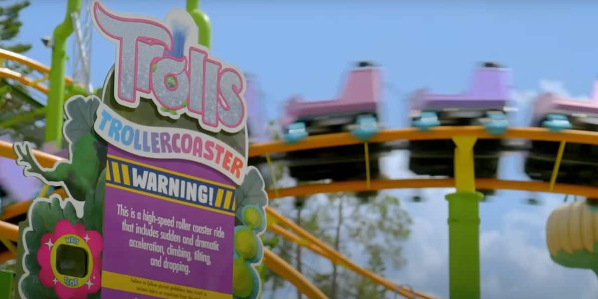 A colorful sign for the “Trolls Trollercoaster” stands in front of a blurred roller coaster ride with pink and blue cars speeding along the yellow track under a blue sky with trees in the background.