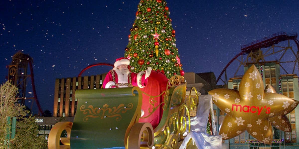 Santa Claus waves from a decorated sleigh in front of a universal, lit Christmas tree at night. Nearby is a giant gold star with the Macy’s logo, with buildings and a roller coaster in the background at Universal Orlando.