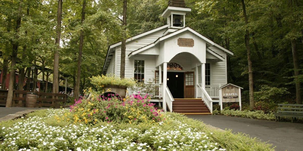 A charming white chapel nestled in lush gardens at Dollywood, surrounded by vibrant blooms and tall trees under sunny skies.