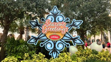 Seasons Greetings sparkles on a giant snowflake sign at Disney Springs, where festive guests mingle amid lush holiday decor.
