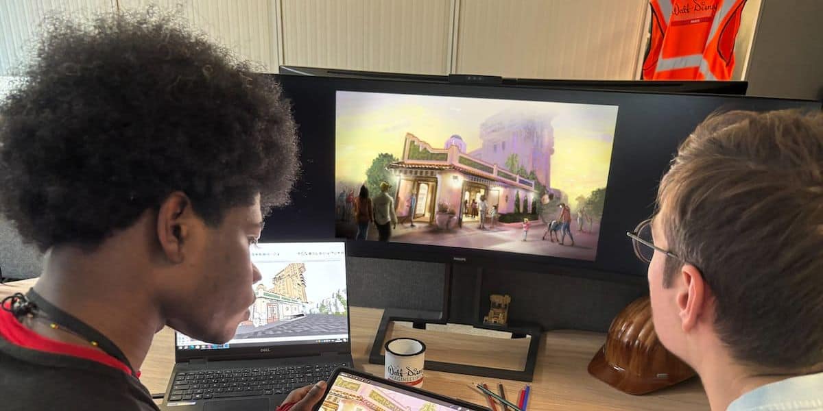 Two designers review a new theme park attraction blueprint on a computer, surrounded by creative planning materials.