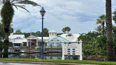 A lively resort with palm trees, a shimmering lake, and signs guiding guests to vacation villas and the grand check-in pavilion.