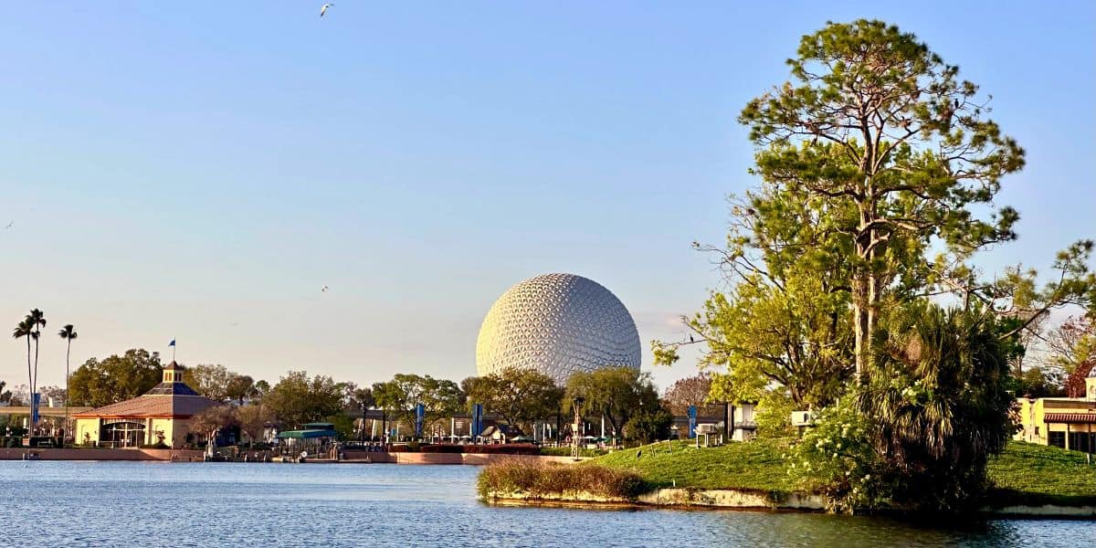 EPCOT’s iconic geodesic sphere towers over World Showcase Lagoon, surrounded by lush trees and park buildings, under a clear sky.