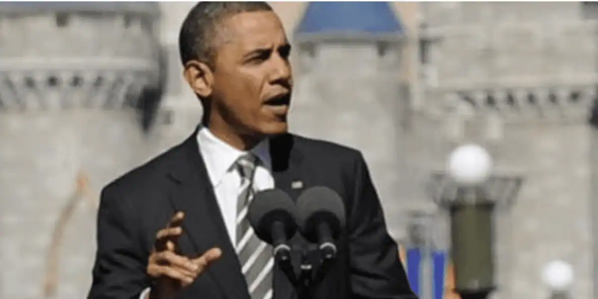 Barack Obama, in a suit, delivers a speech at microphones—just like his famous Disney World animatronic once did.