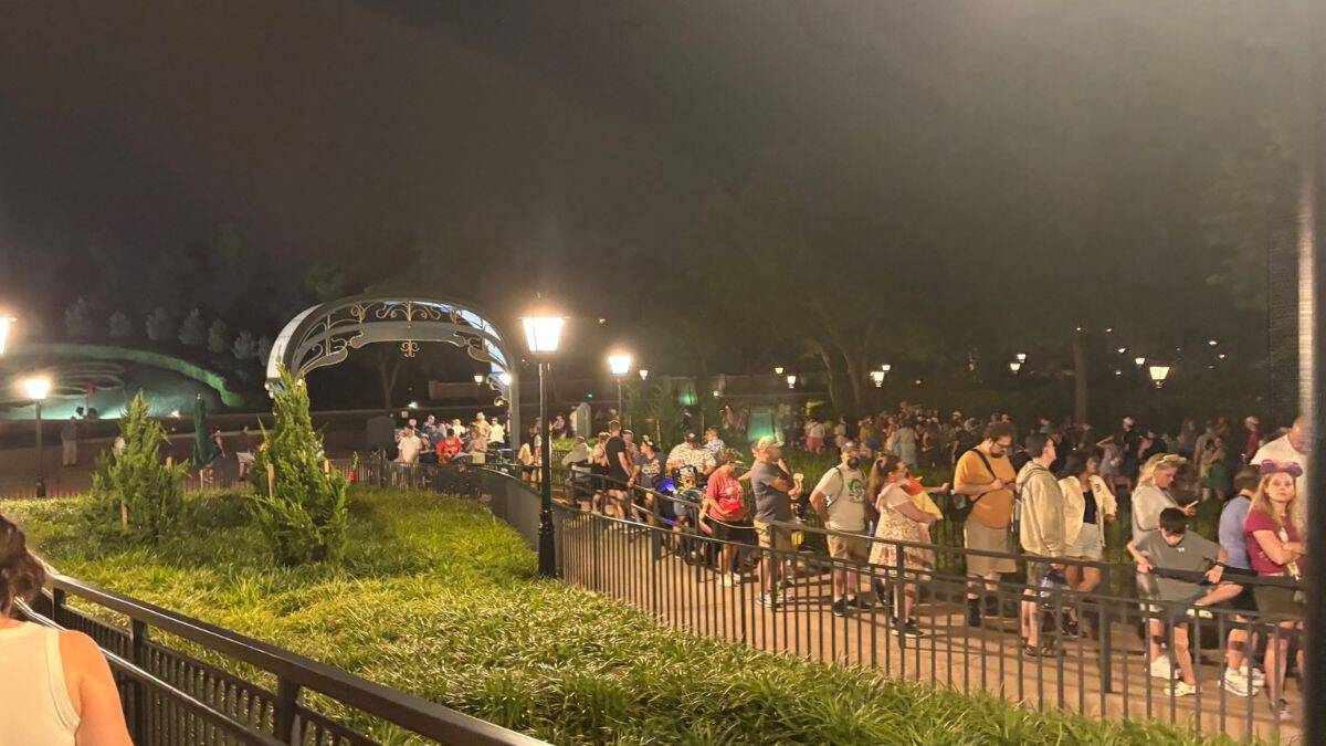 Guests form a winding nighttime queue by a glowing arch and lush gardens, evoking the iconic lines at Disney World transit.