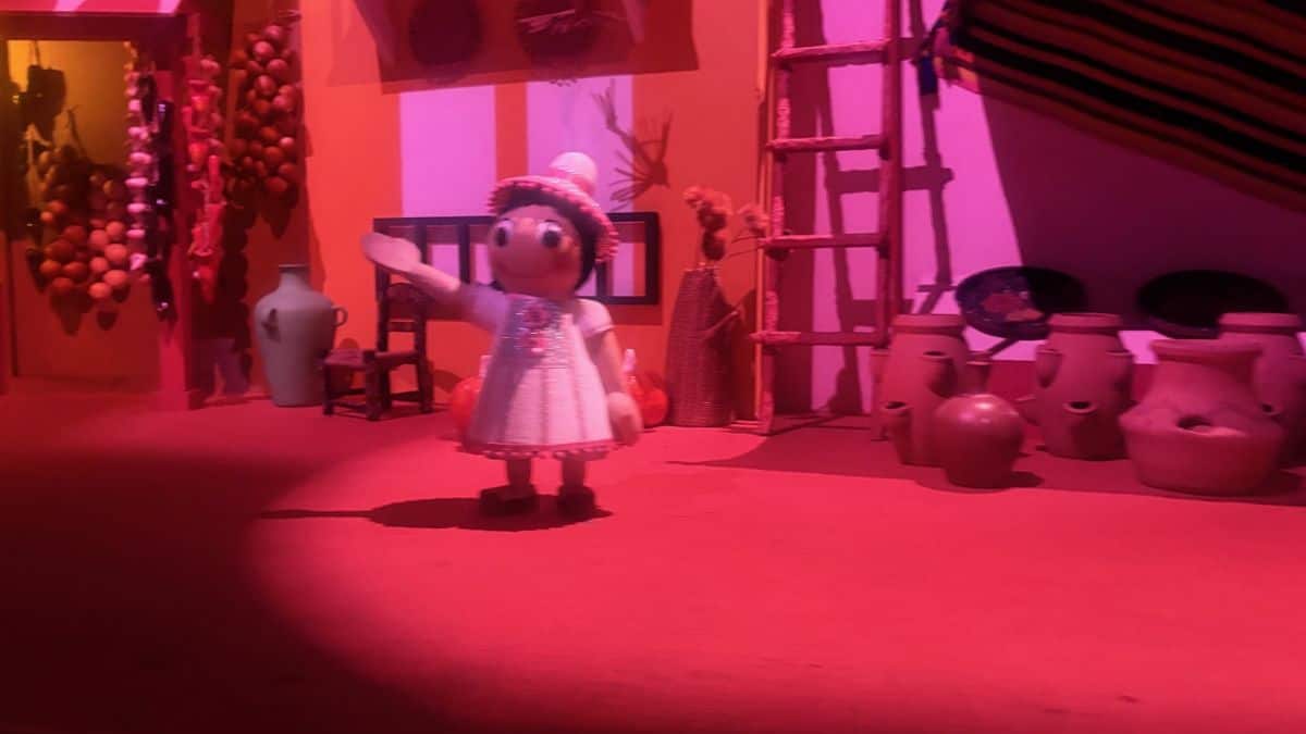A Small World-style doll in vibrant dress pops amid bright pink lights and lively clay pots, all fresh and enchantingly new.