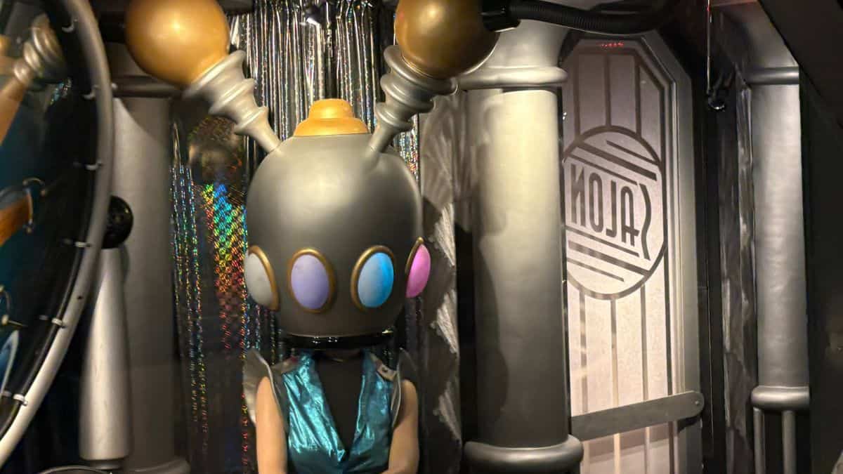 The Futuristic Hair Salon Patron, sporting a robot helmet with antennae, greets guests in Tomorrowland’s metallic "SALON" scene.