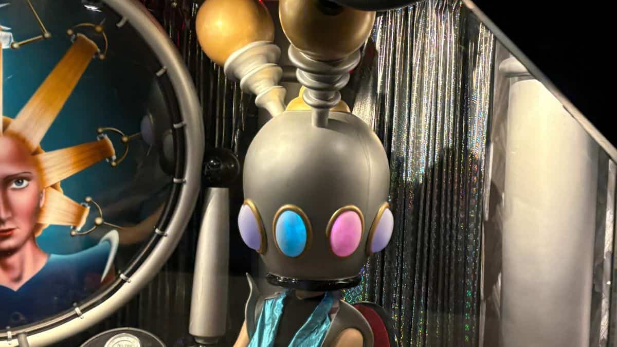 Futuristic Hair Salon Patron with rainbow lights on its head sits by metallic curtains, evoking PeopleMover’s Tomorrowland vibes.