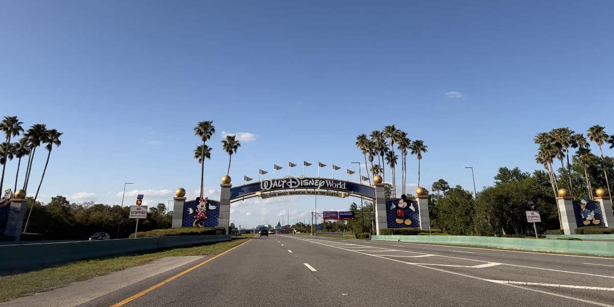 Entrance road lined with palm trees leading up to the iconic Walt Disney World arch under a bright, sunny sky.