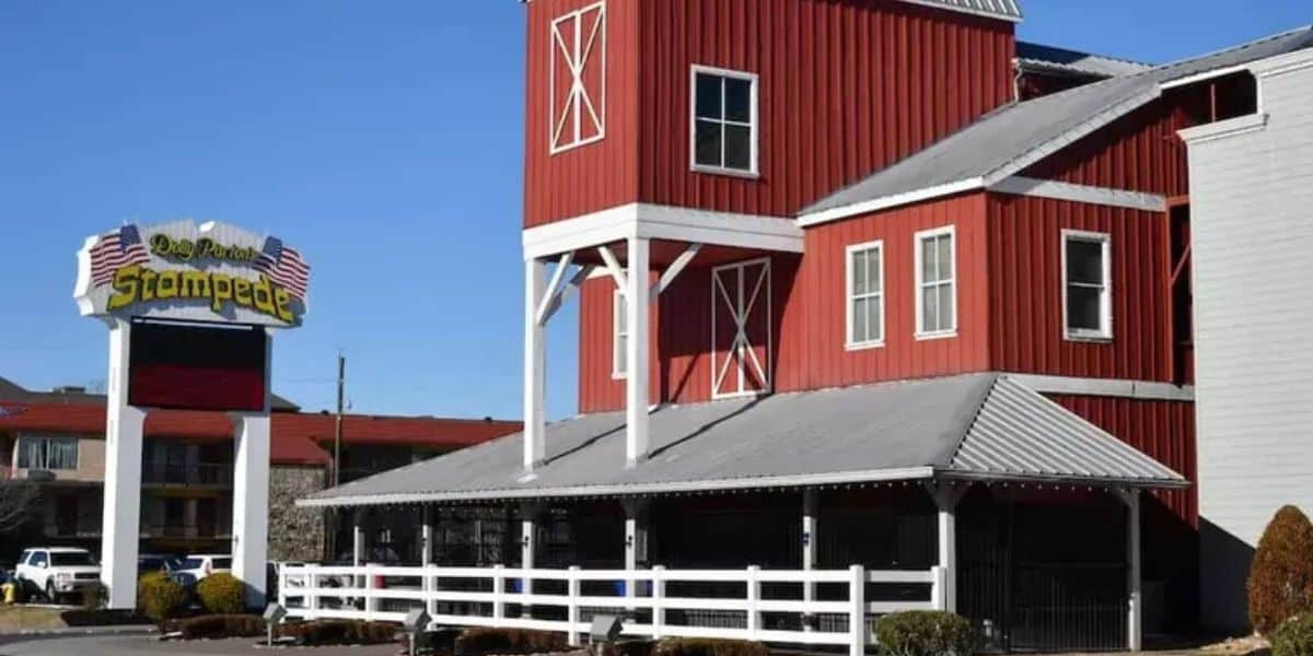 The Stampede barn, with its iconic red exterior and white fence, welcomes guests under a bright sun at the entrance.