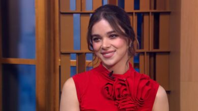 A performer in a vibrant red floral dress beams while being interviewed on a themed entertainment news set.
