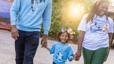 Guests in Bluey shirts stroll hand-in-hand under the sun, beaming with excitement for their Disney Bluey Experience adventure.