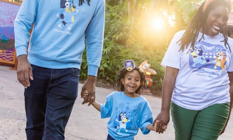 Guests in Bluey shirts stroll hand-in-hand under the sun, beaming with excitement for their Disney Bluey Experience adventure.