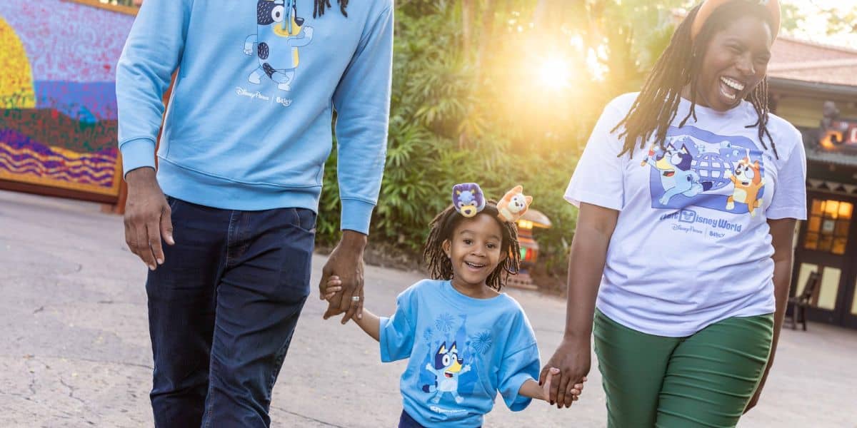 Guests in Bluey shirts stroll hand-in-hand under the sun, beaming with excitement for their Disney Bluey Experience adventure.
