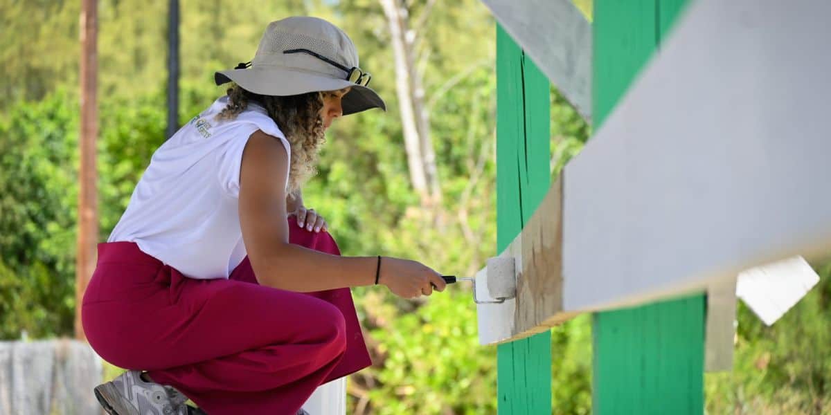 A costumed entertainer in a themed hat and red pants adds fresh paint to a wooden fence at an outdoor park attraction.