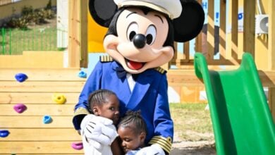 Mickey Mouse, dressed as a captain, warmly hugs two kids by a playground slide at Disney World.