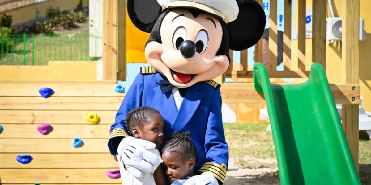 Mickey Mouse, dressed as a captain, warmly hugs two kids by a playground slide at Disney World.