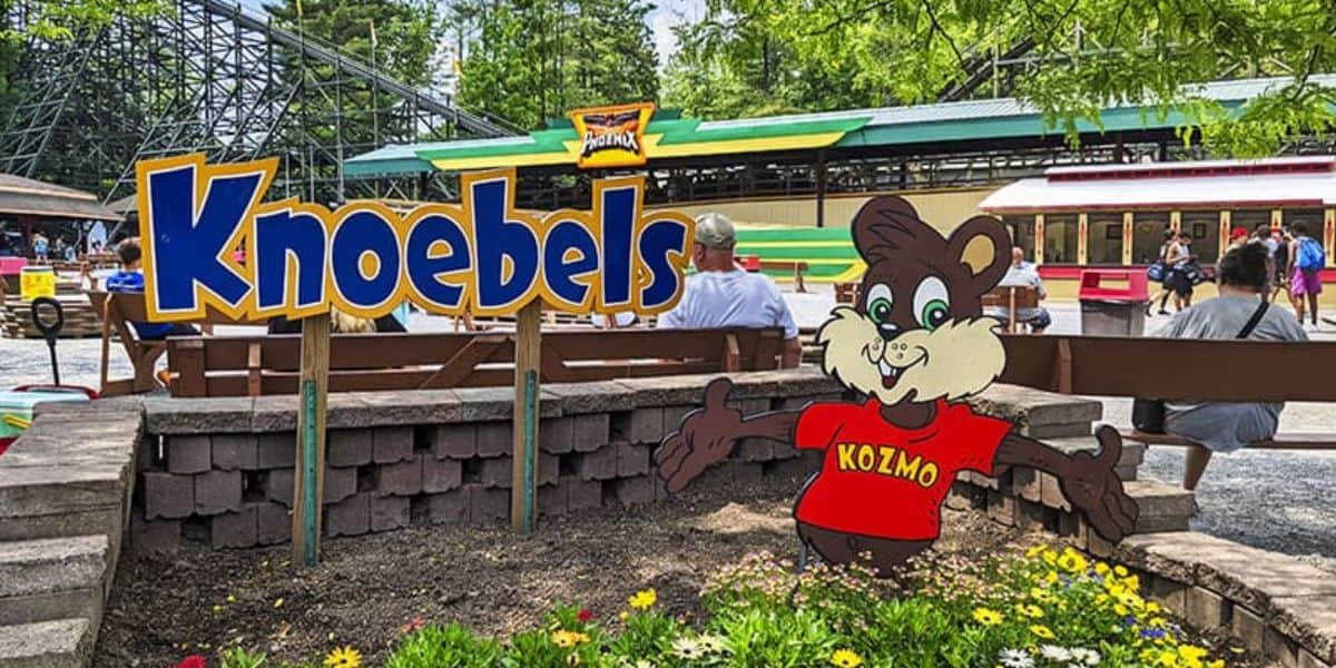 Kozmo greets guests under the Knoebels sign, surrounded by benches, vibrant flowers, and classic park rides in the background.