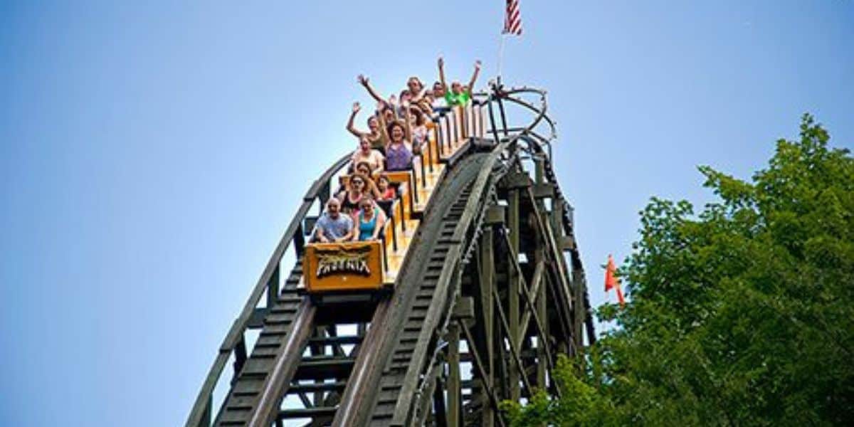 Thrill-seekers with hands up race down the classic Timber Tornado coaster’s steep drop, blue skies and forest scenery all around.