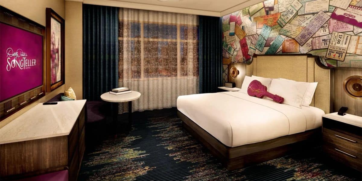 A Hard Rock Hotel room with a king bed, guitar pillow, concert ticket wallpaper, flat-screen TV, desk, and window with drapes.