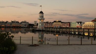 A lively boardwalk at Disney Springs, featuring a lighthouse, waterfront dining, and glowing sunset reflections on calm water.