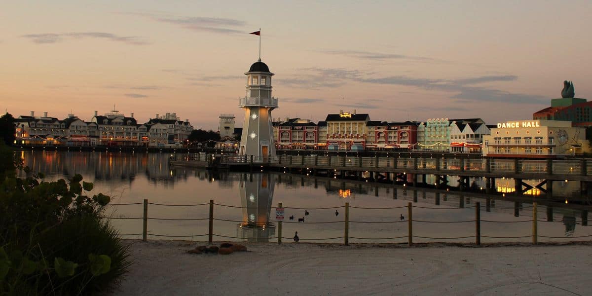 A lively boardwalk at Disney Springs, featuring a lighthouse, waterfront dining, and glowing sunset reflections on calm water.