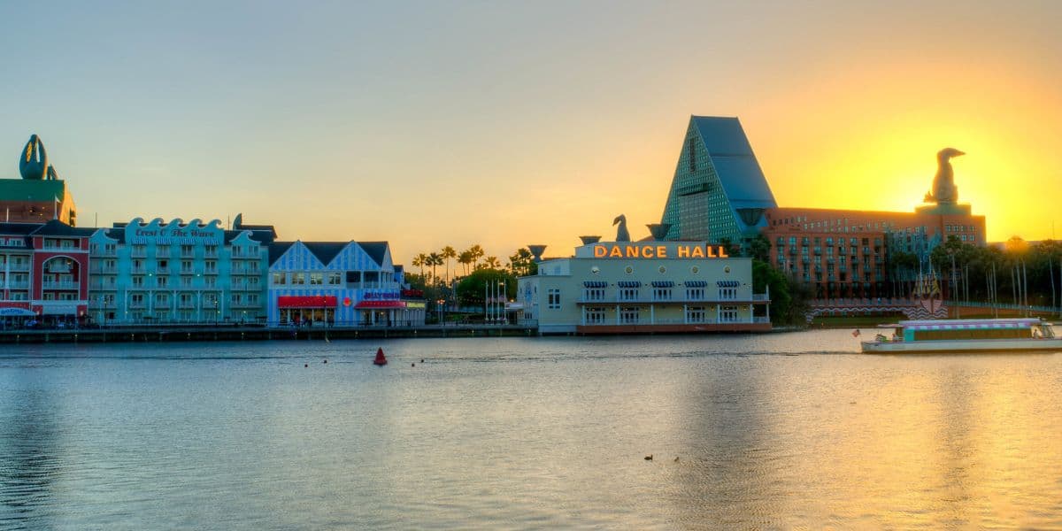 Sunset over Disney Springs with its vibrant waterfront, entertainment venues, and a boat cruising past lively themed buildings.