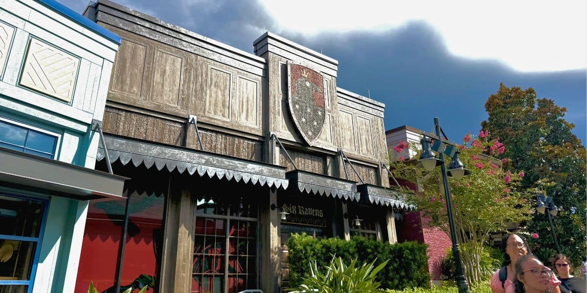 Guests stroll by a shield-decorated restaurant in Disney Springs, framed by lush summer plants and cloudy Florida skies.