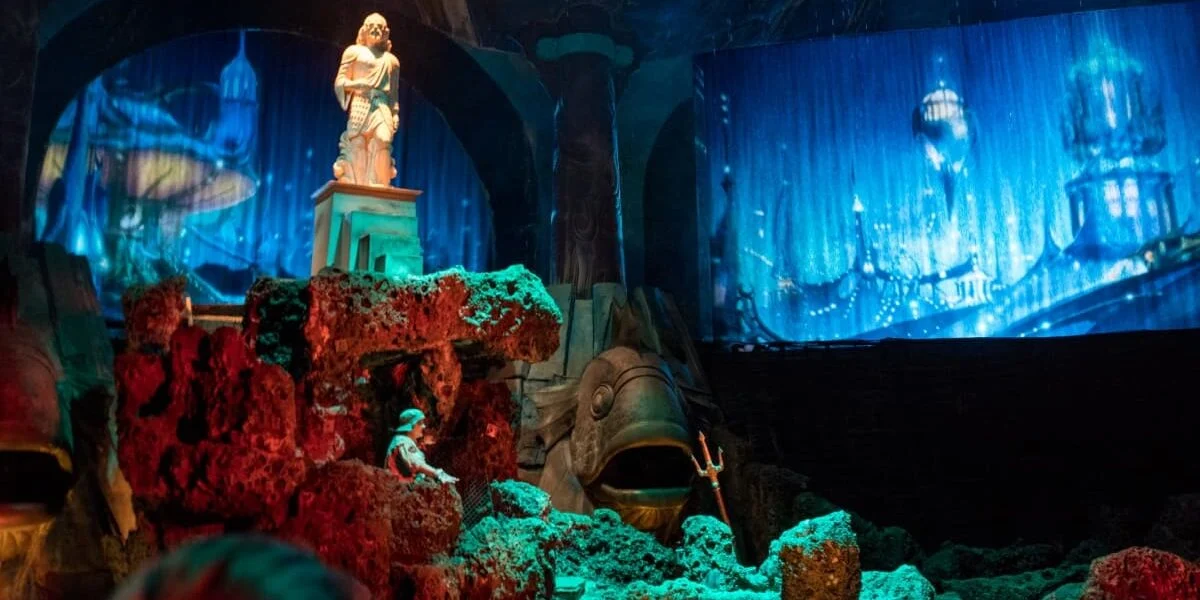 Journey into a mesmerizing underwater world at the park’s immersive Atlantis zone, featuring a majestic statue centerpiece surrounded by vibrant rocks, an impressive animatronic fish gliding through crystal-blue waters, and a dazzling city skyline illuminated in deep ocean hues.