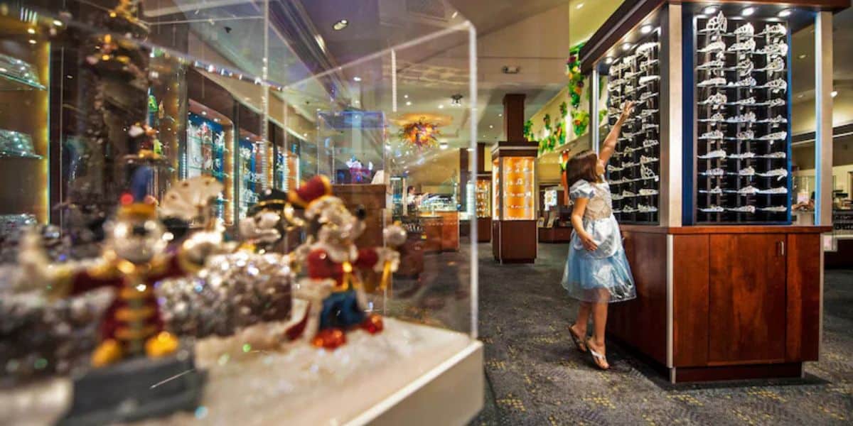 A young guest in a Cinderella-inspired blue dress excitedly reaches for souvenirs inside a vibrant, well-lit gift shop with gleaming glass display cases at the theme park.