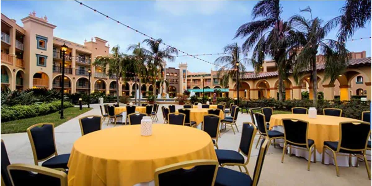 Outdoor resort tables covered in bright yellow cloths sit beneath swaying palm trees and glowing string lights, setting a festive scene that captures the enchanting, celebratory spirit of Disney World in August.