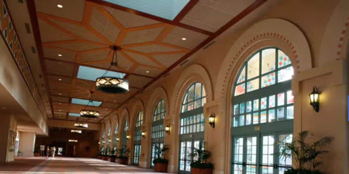 Grand, sunlit hallway lined with soaring arched windows and lush potted plants, featuring a lavishly detailed ceiling and signature Disney hanging lights—capturing the upscale magic of a Disney World resort lobby in its final moments before closing on August 10.