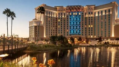 A vibrant Disney resort hotel illuminated at dusk, its lights shimmering on a serene lagoon, with iconic palm trees and colorful gardens welcoming guests to the magical atmosphere.
