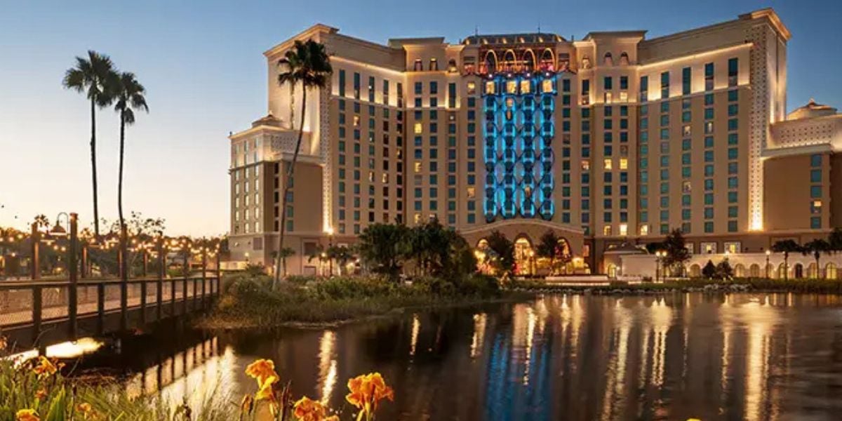 A vibrant Disney resort hotel illuminated at dusk, its lights shimmering on a serene lagoon, with iconic palm trees and colorful gardens welcoming guests to the magical atmosphere.